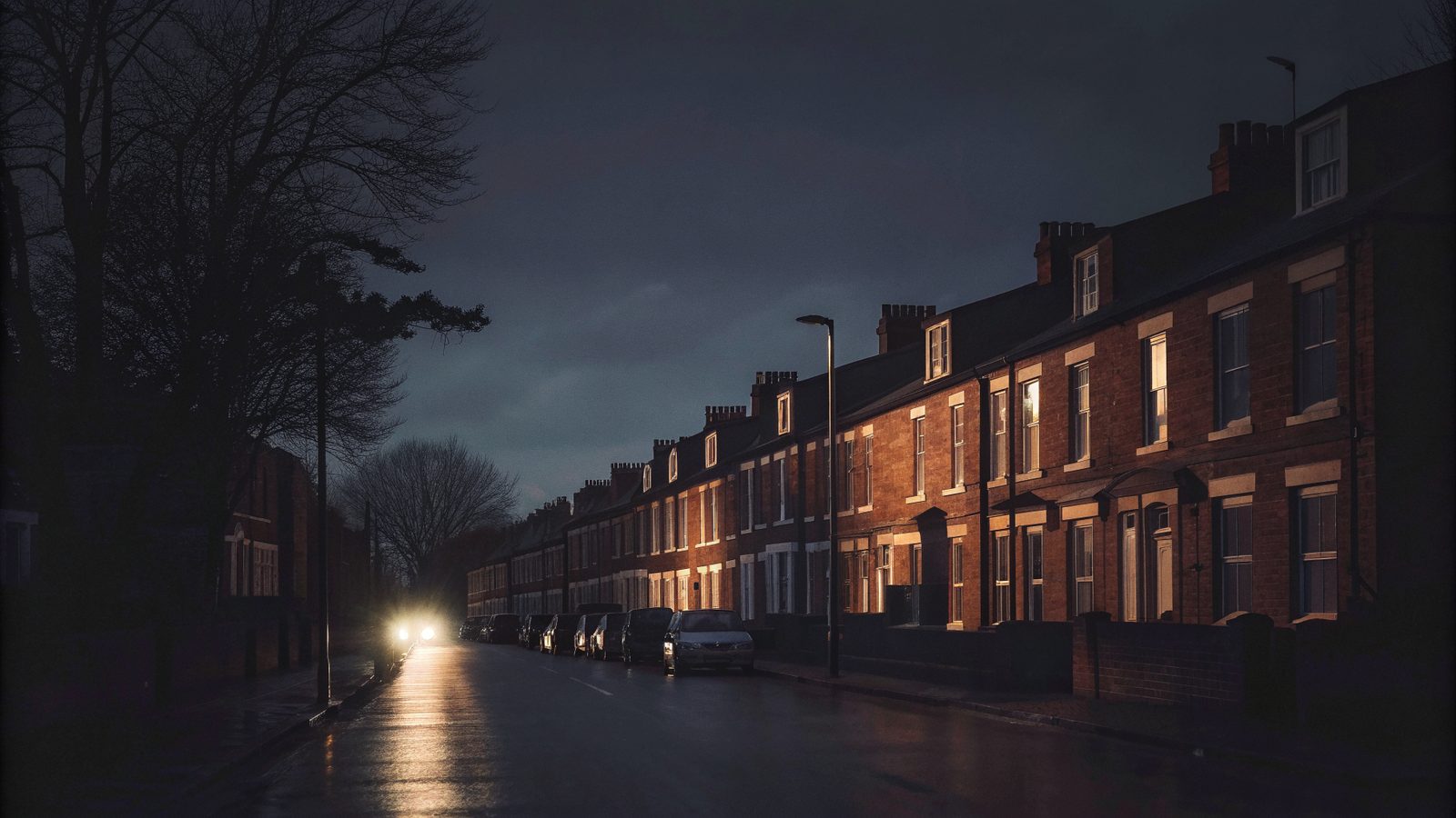 A long row of red brick terraced houses on a tree-lined street at dusk, warm light in a few windows and one car's headlights on the wet road